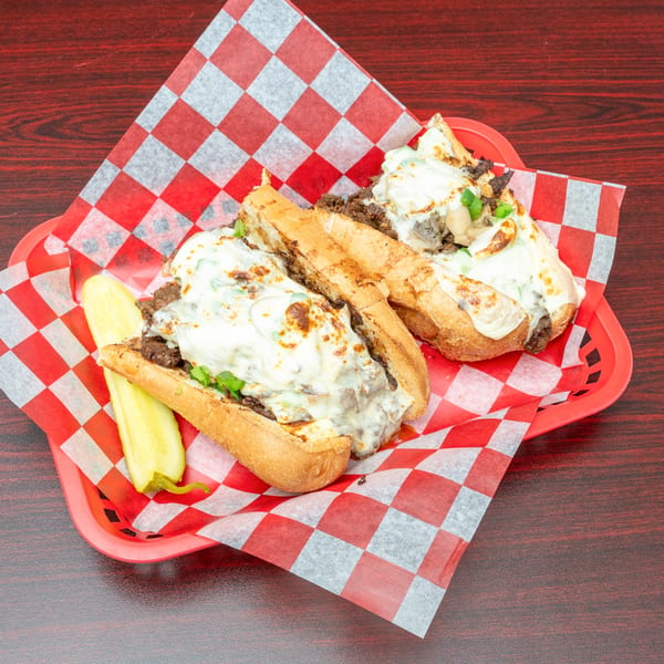 Two cheesy steak and mushroom subs with a pickle spear, served in a red basket with checkered paper.