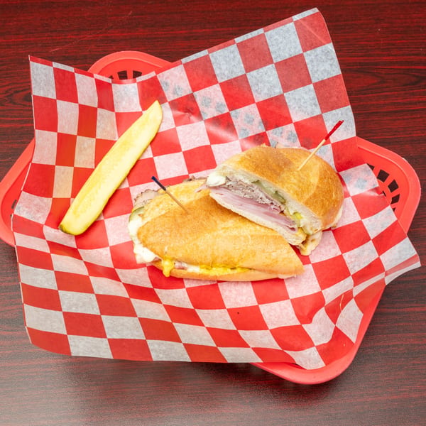Cuban sandwich with ham, pork, cheese, mustard, and pickles, served on red checkered paper.