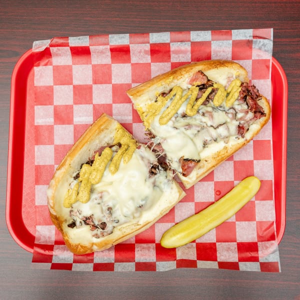 Cheesesteak sandwich cut in half, topped with mustard, served with a pickle on a red checkered tray.