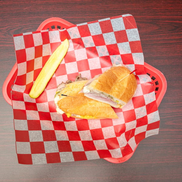 Cuban sandwich with a pickle spear, served in a red basket with checkered paper.