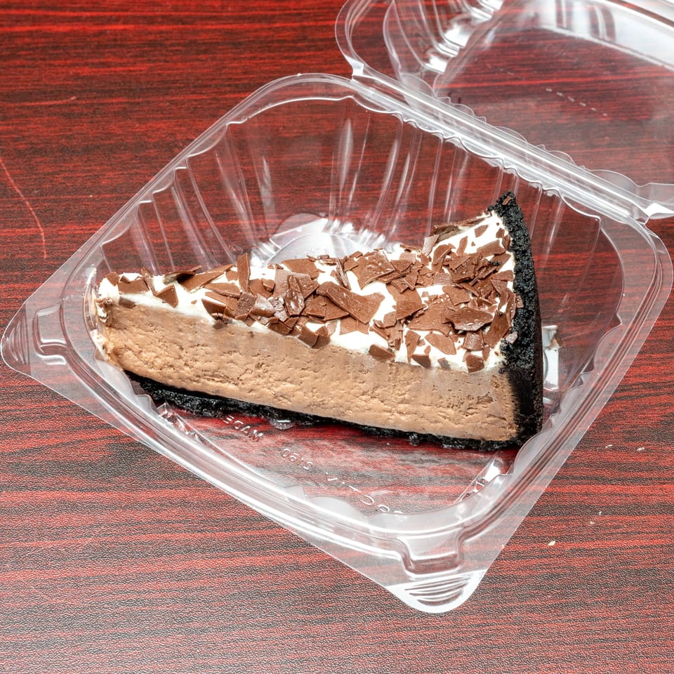 Slice of chocolate cheesecake with whipped cream and chocolate shavings in a clear plastic container.