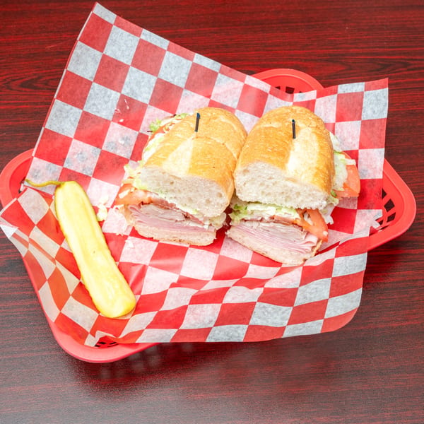 A large deli sub sandwich, cut in half, with a pickle spear in a red basket with checkered paper.