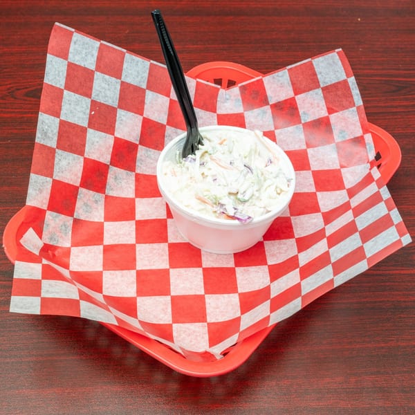 Creamy coleslaw in a white cup with a black fork, served in a red checkered basket.
